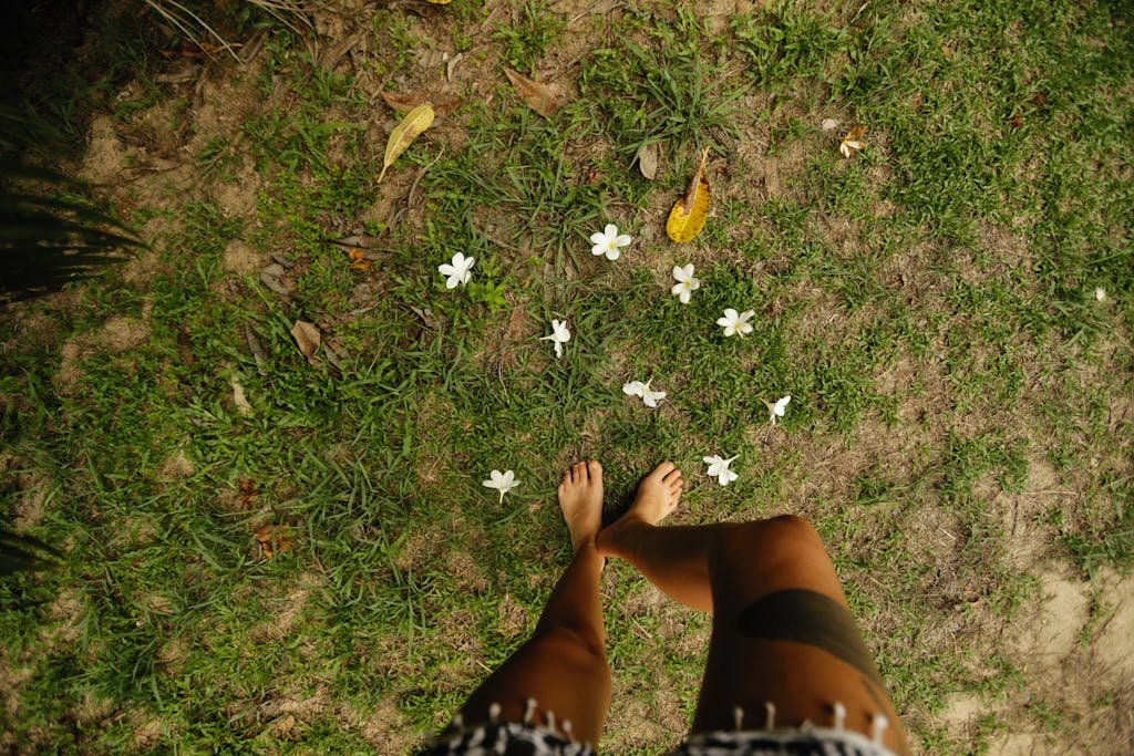 A relaxing top view of a barefoot person standing on grass surrounded by white Plumeria flowers.