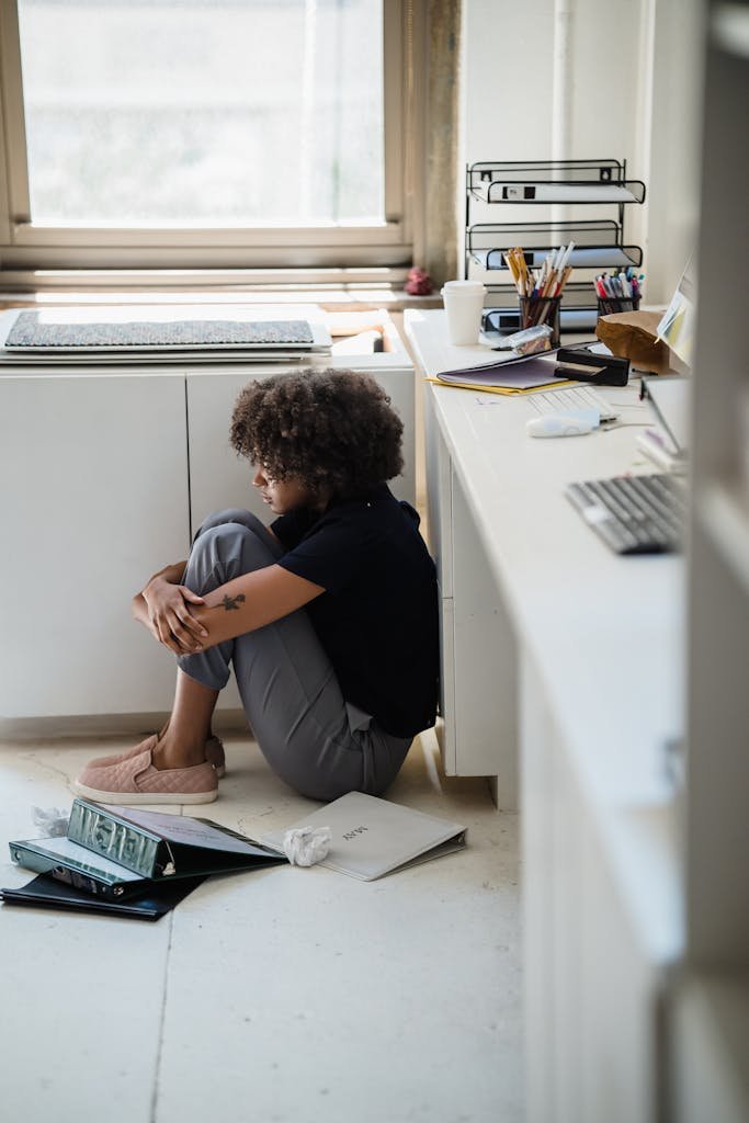A woman sits stressed on the floor of an office, surrounded by papers.