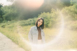 A young woman wearing a jean jacket stands in a sunlit rural setting, exuding style and confidence.
