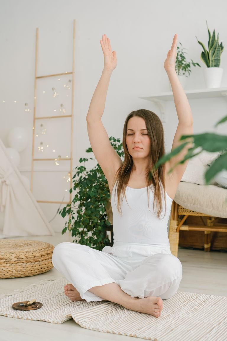 A woman sits in a living room meditating with arms raised, surrounded by plants.