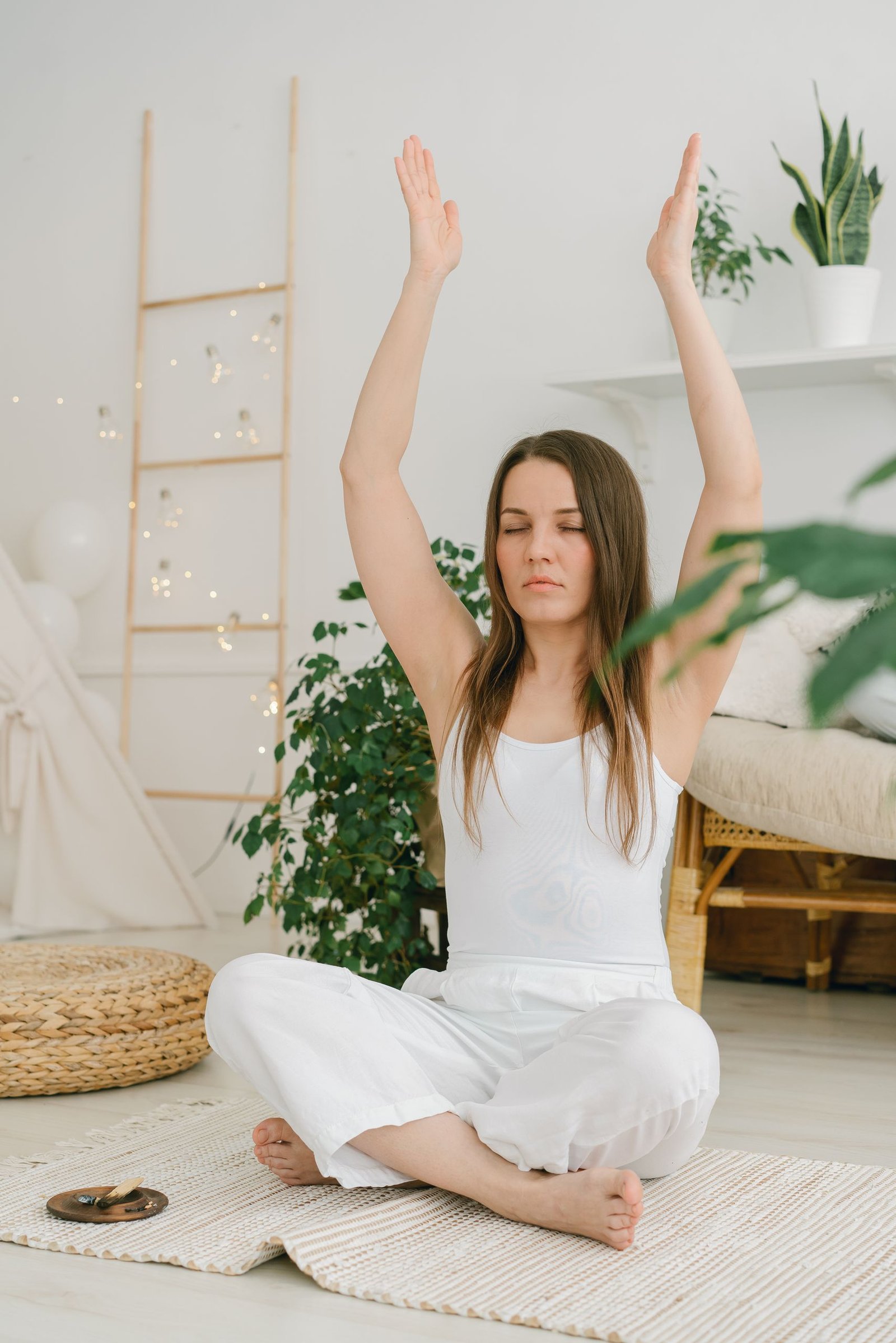 A woman sits in a living room meditating with arms raised, surrounded by plants.