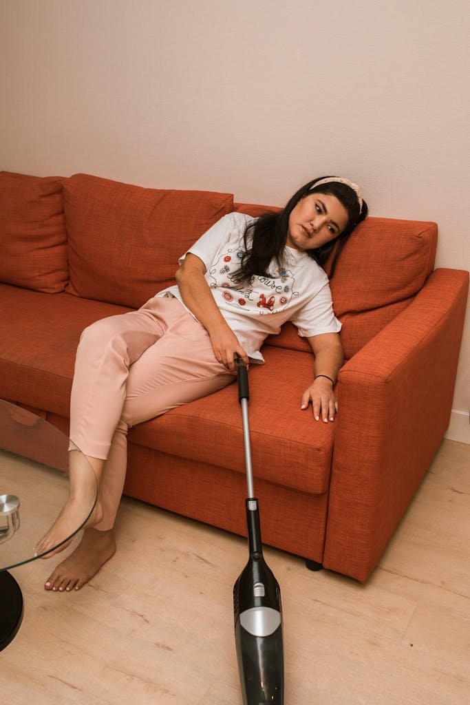 Asian woman lounging on a sofa with a vacuum cleaner, looking tired and relaxed.