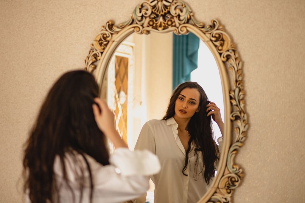 Elegant woman with long brunette hair gazing at her reflection in an ornate mirror, wearing white shirt indoors.