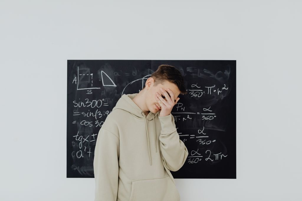 Teen boy covers face in frustration in front of a chalkboard filled with math equations, reflecting academic stress.